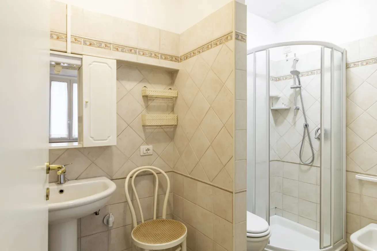 A tiled bathroom with a sink, shower enclosure, and chair.