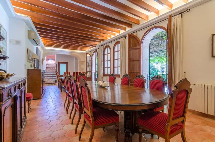 A dining room with a long table and red upholstered seating beneath exposed wooden ceiling beams.