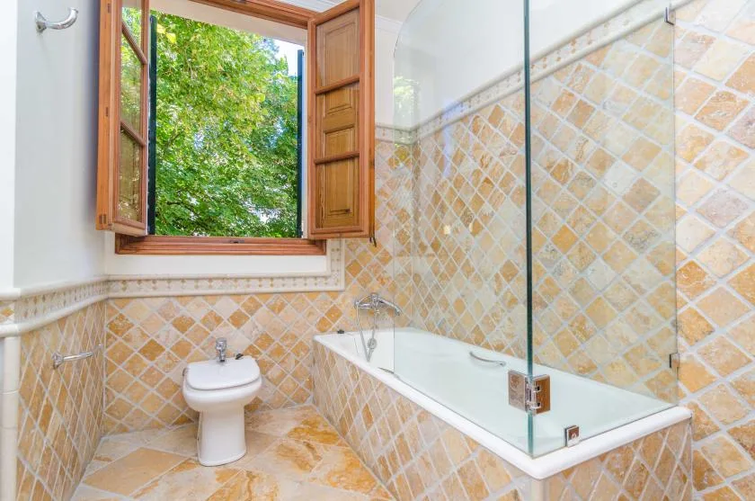 A tiled bathroom with a bathtub, glass screen, and open window.