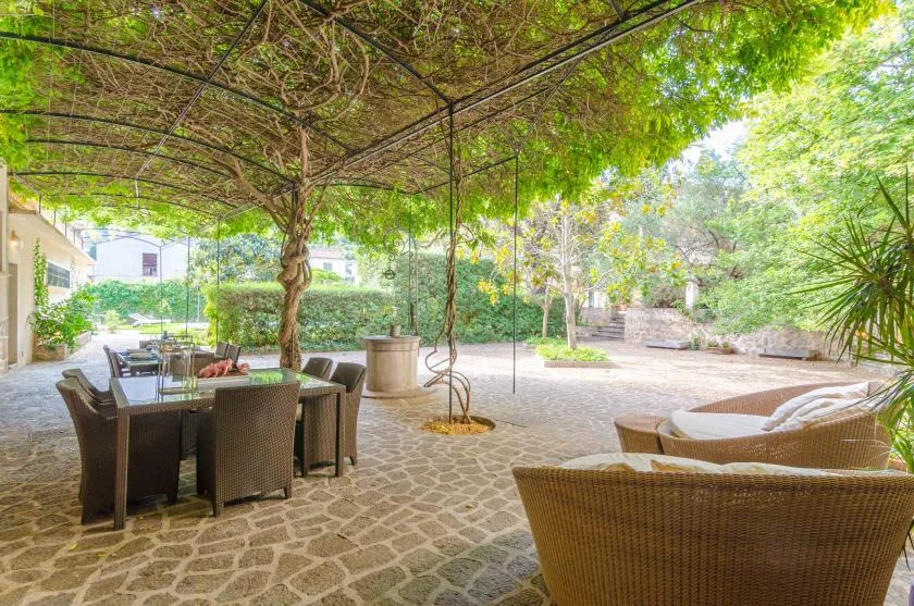 A leafy outdoor patio has a dining table and lounge seating beneath a vine-covered canopy.