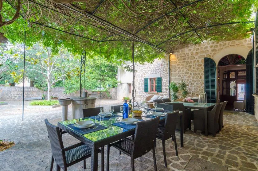 A shaded outdoor dining area with tables beneath a leafy canopy beside a stone building.