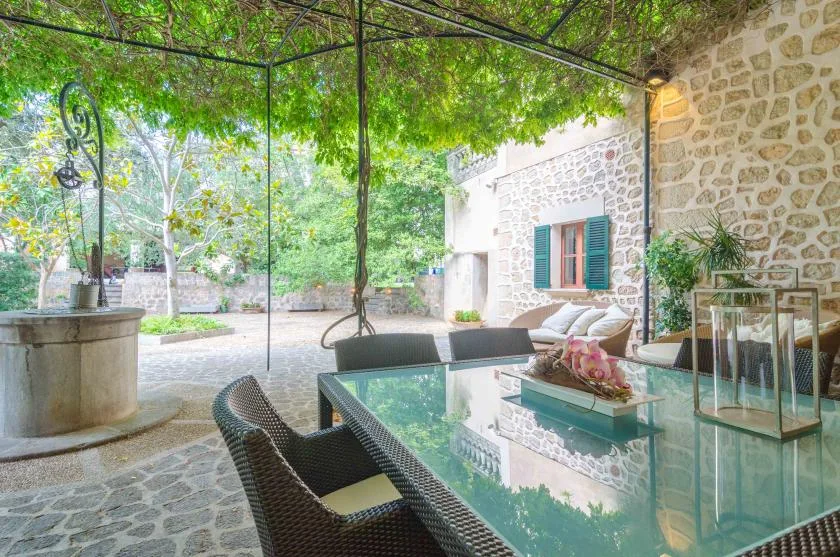 A covered stone patio with outdoor seating, a glass table, and surrounding greenery.