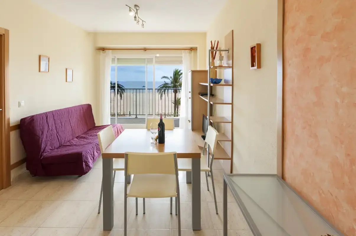 A living and dining area with a table, seating, and a balcony overlooking the sea.