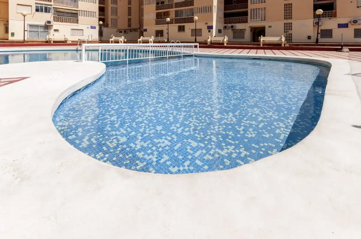 An outdoor swimming pool with blue tiled water and a surrounding paved deck.