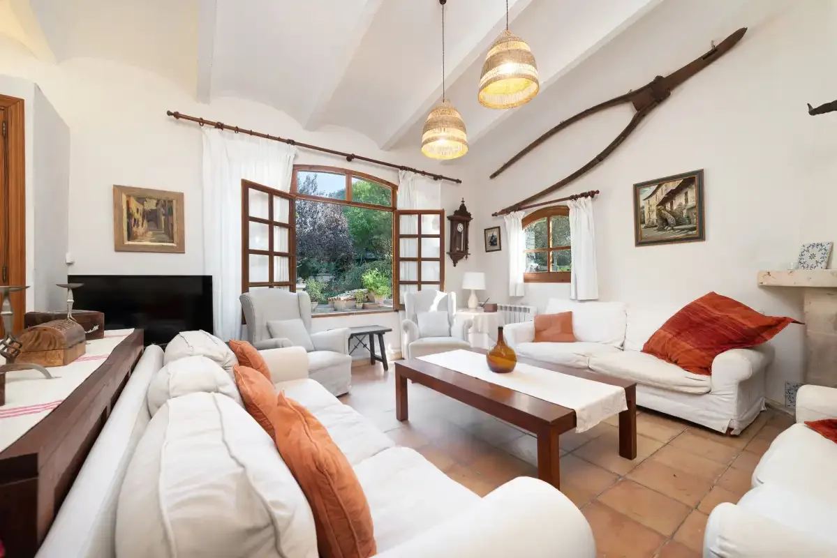 Una sala de estar luminosa con asientos blancos, una mesa de centro de madera y ventanas abiertas.