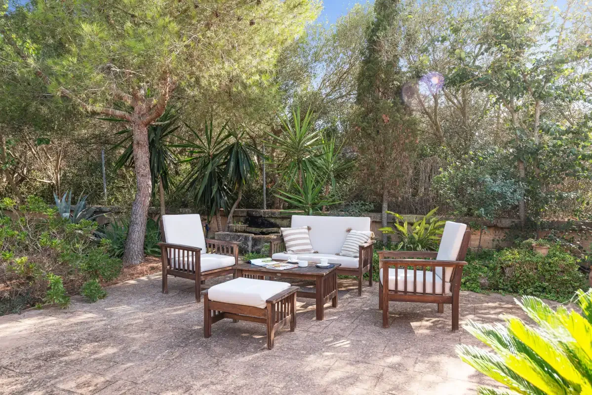Una zona de estar al aire libre con muebles de madera está rodeada de árboles y plantas.