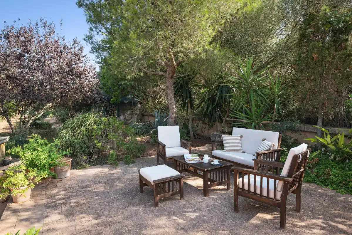 Un patio ajardinado con asientos al aire libre y una mesa baja bajo árboles frondosos.