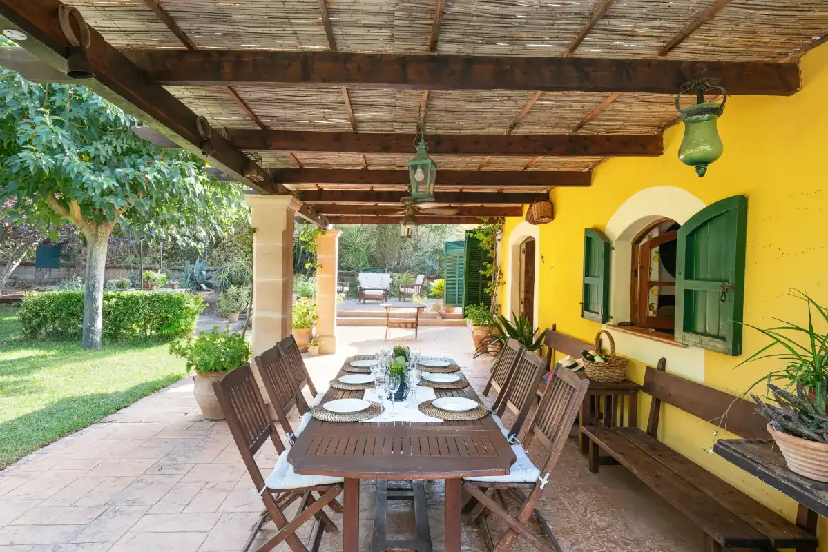 Un comedor exterior cubierto tiene una mesa larga junto a paredes amarillas y contraventanas verdes.