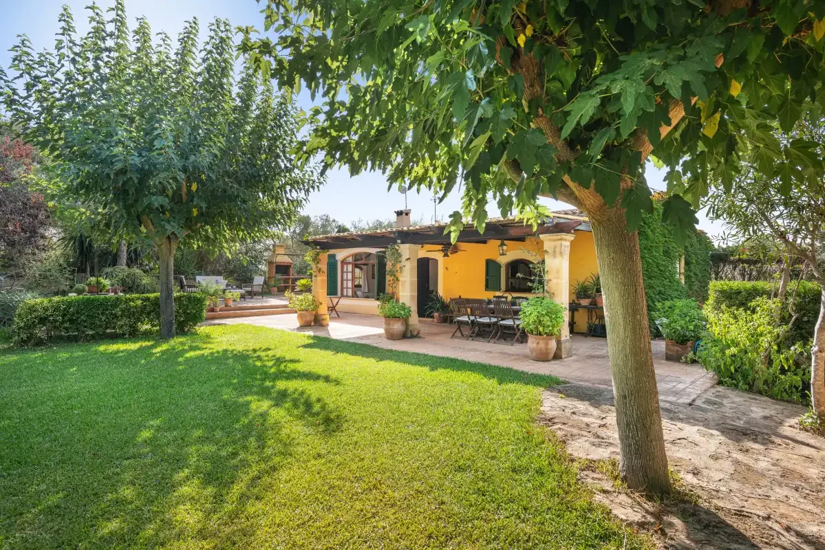 Una casa amarilla da a un jardín con césped, árboles y un patio pavimentado.
