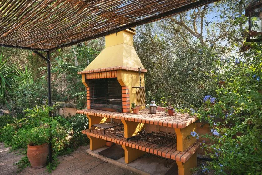 Una parrilla de ladrillo al aire libre está bajo un toldo de listones en un jardín.