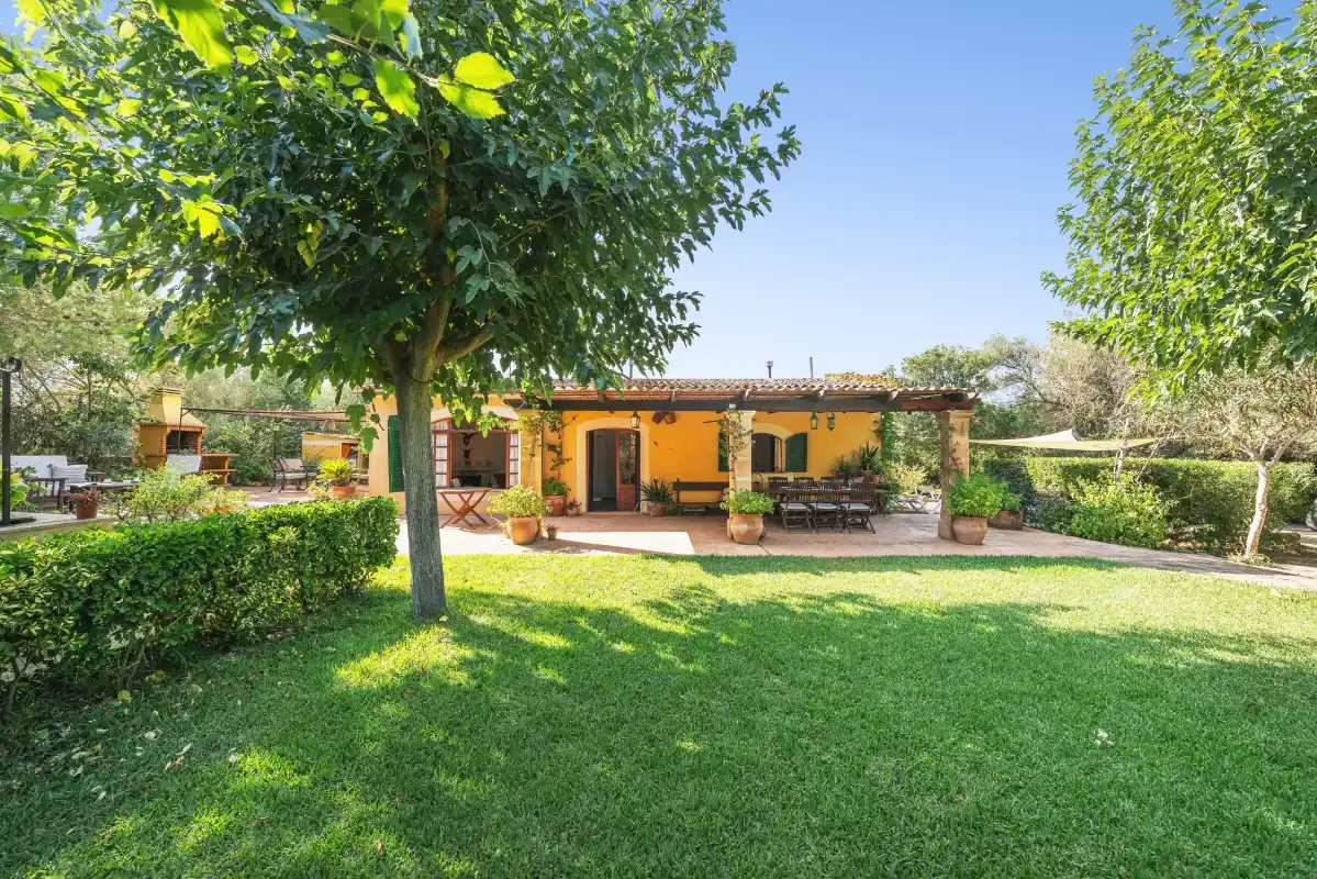 Un césped de jardín y árboles frondosos conducen a una casa de una planta con un patio sombreado.