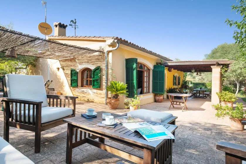 Un patio soleado de piedra con asientos al aire libre junto a una casa con contraventanas verdes.