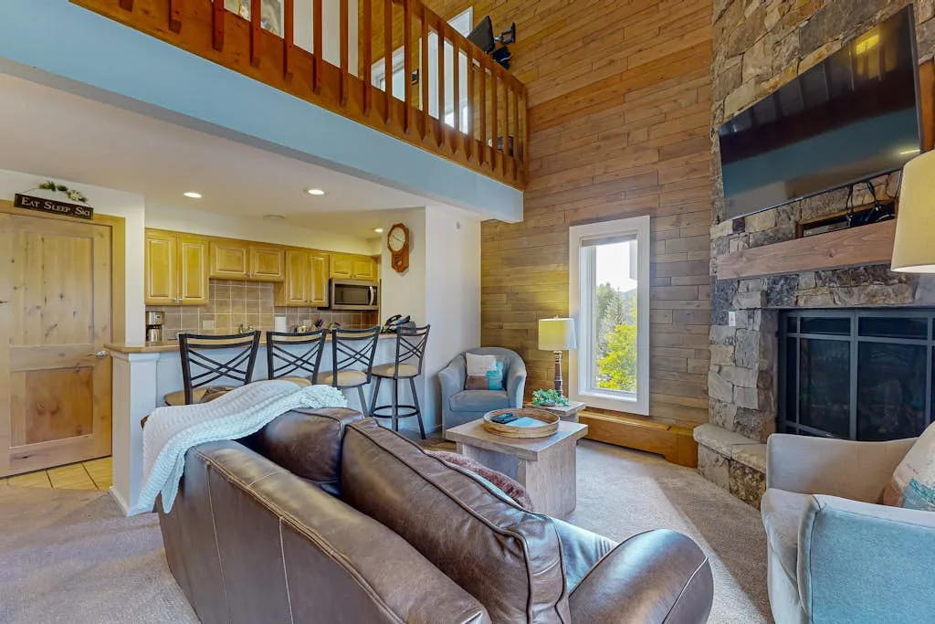 Una sala con paredes de madera se abre a una cocina, con un balcón elevado arriba.