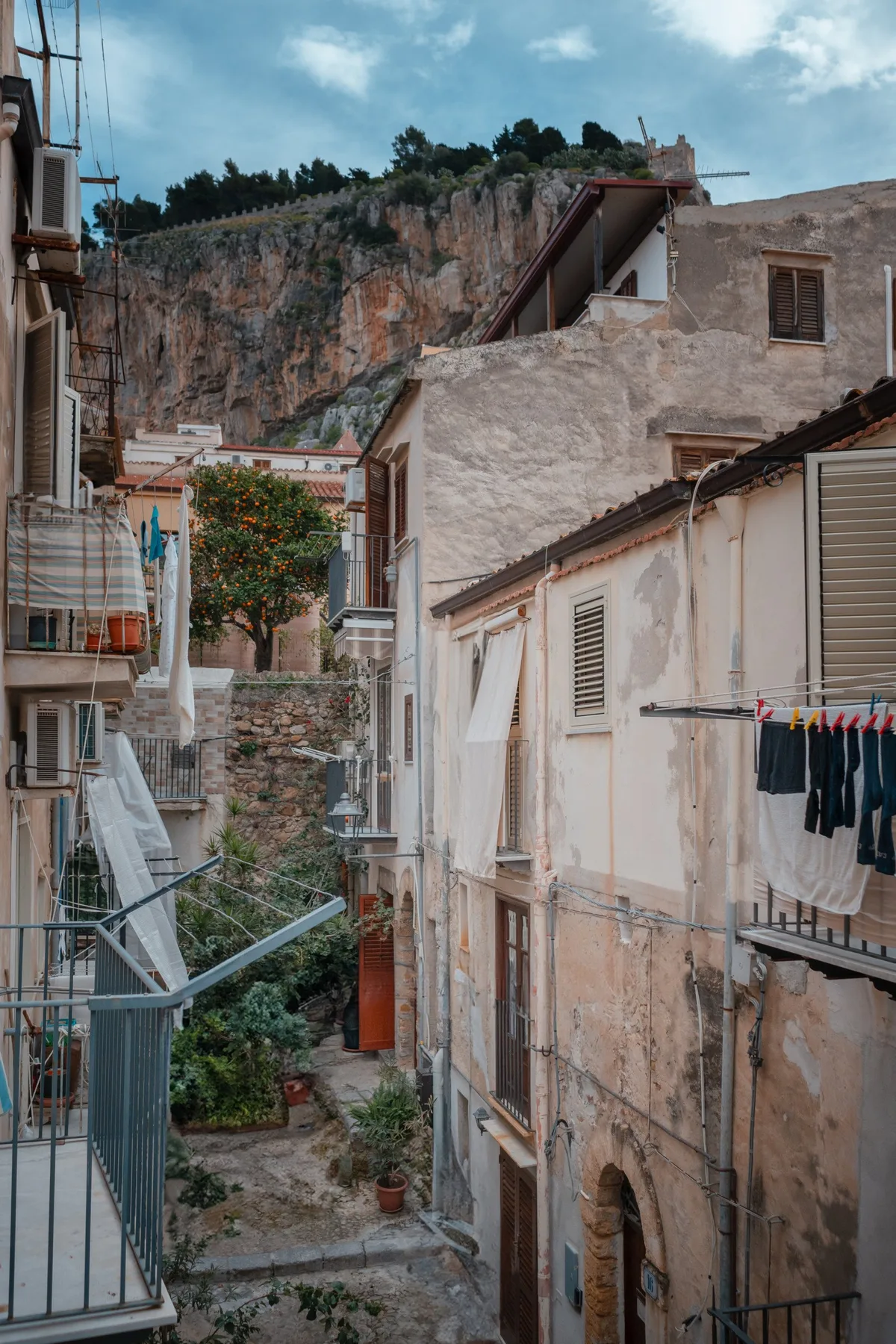 A narrow residential lane runs between closely spaced buildings below a rocky hillside.