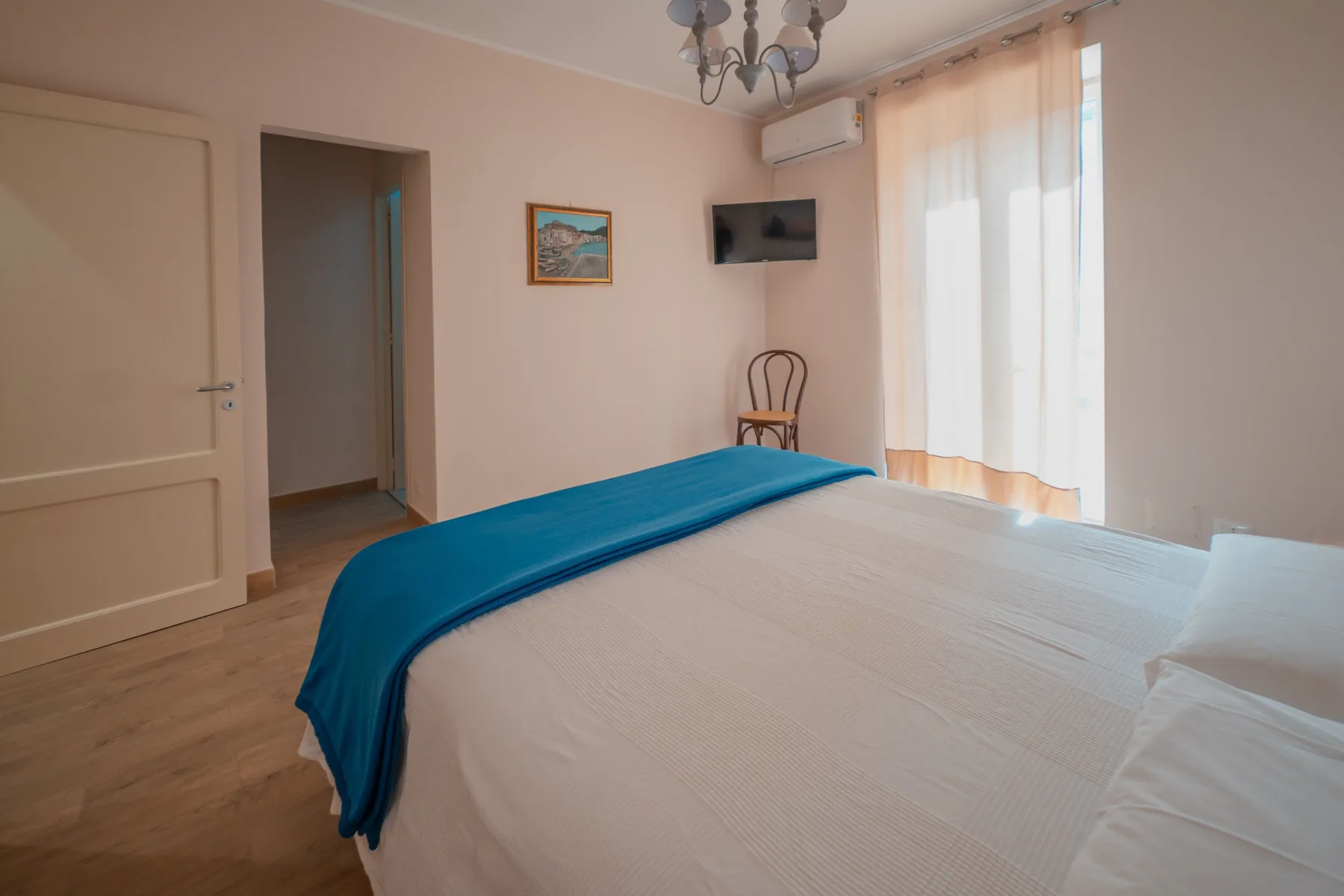 A bedroom with a bed covered by white bedding and a blue runner beside a curtained window.