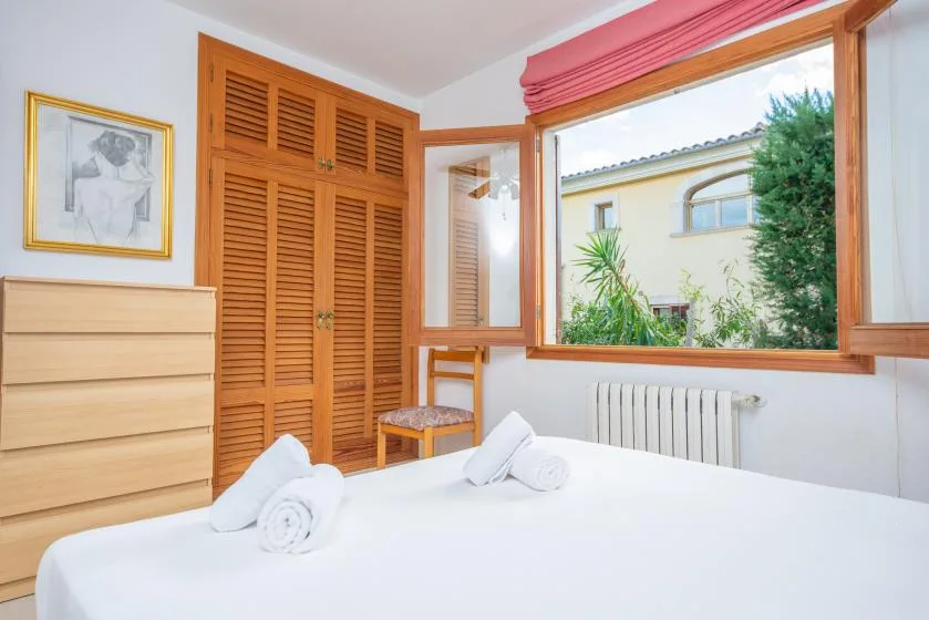 A bedroom with white bedding, wooden storage, and a window overlooking greenery.
