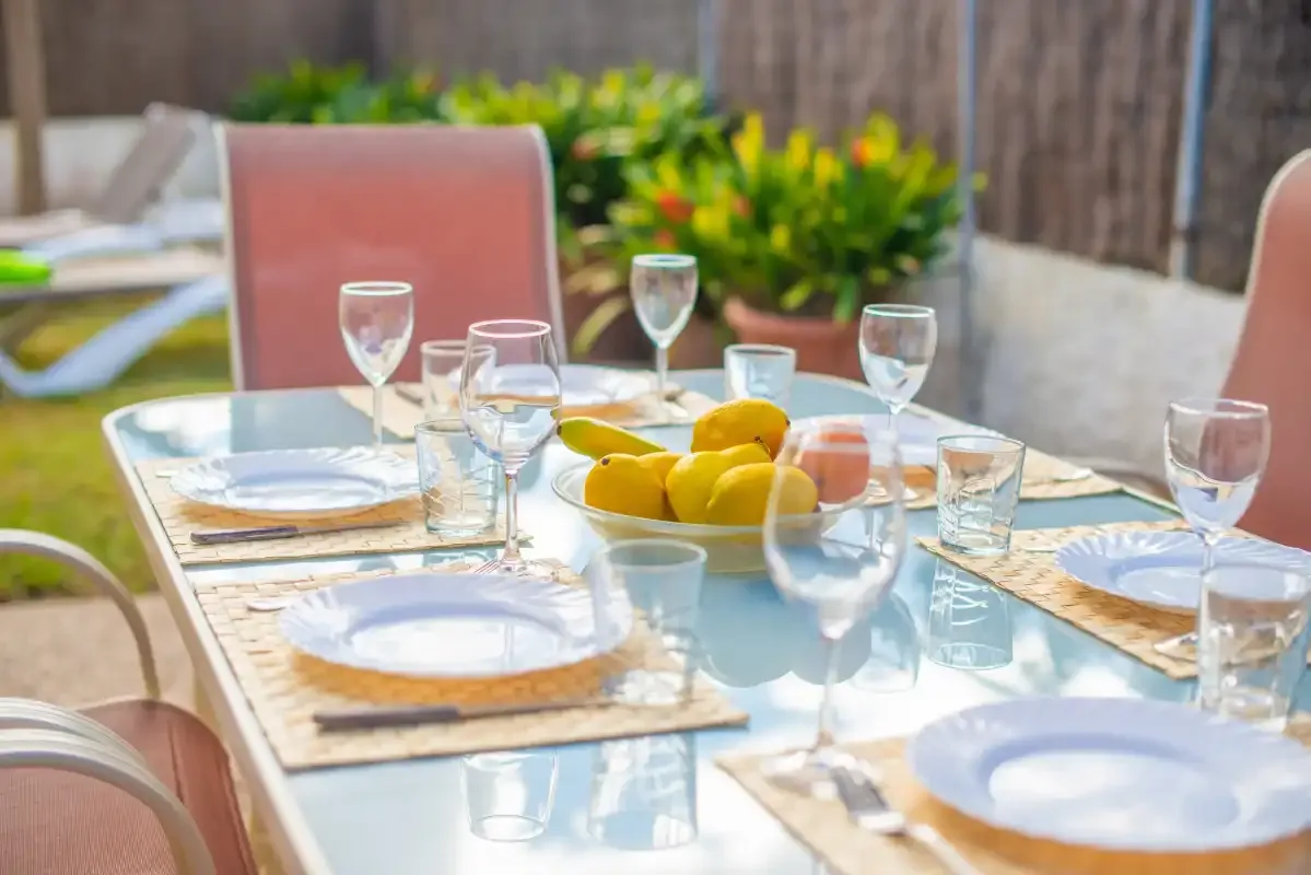 An outdoor dining table is set with plates, glasses, and a bowl of fruit.