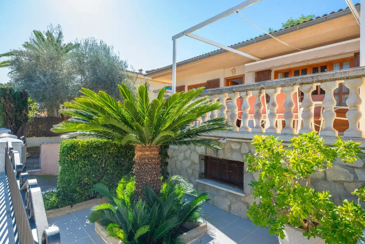 A sunny outdoor patio beside a stone terrace, with leafy plants and a balcony above.