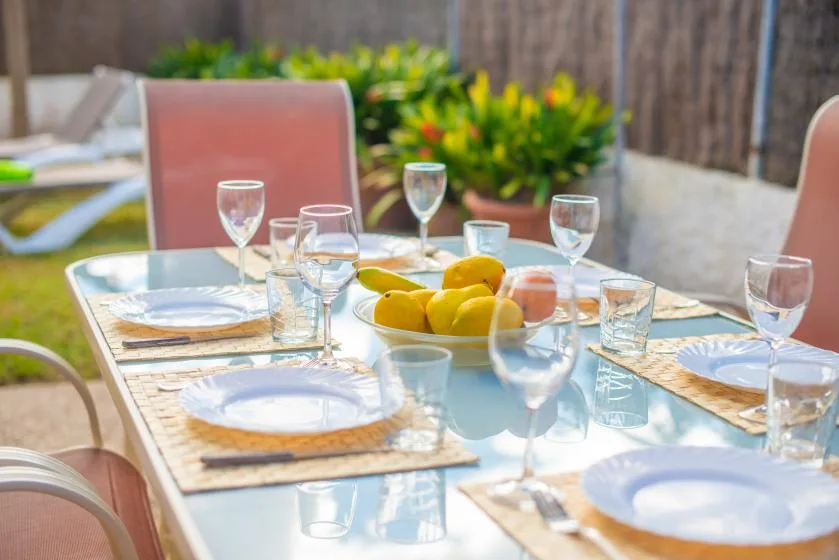 An outdoor dining table is set with plates, glasses, and a bowl of fruit.
