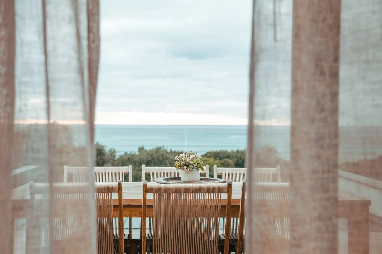 A dining table and chairs stand by a window overlooking the sea.