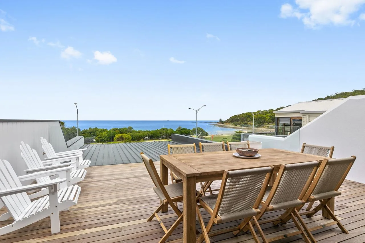 Una mesa de comedor de madera y asientos al aire libre miran al mar desde una terraza.