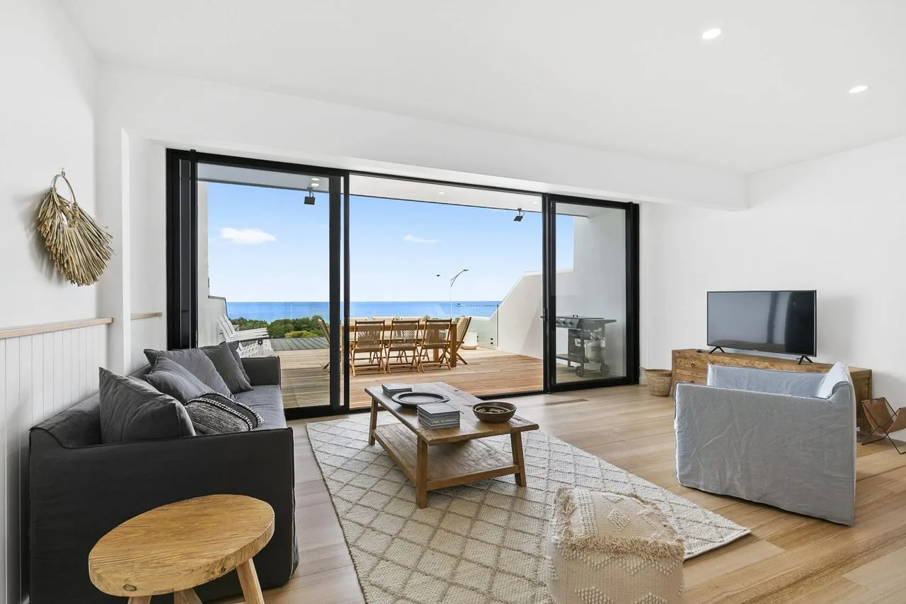 A bright living room opens onto a deck with outdoor seating and a sea view.
