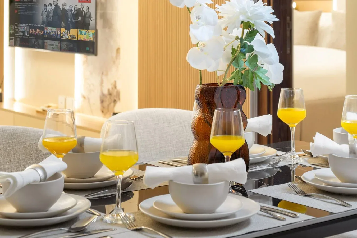 Una mesa de comedor está preparada con vajilla blanca, bebidas amarillas y un jarrón con flores.