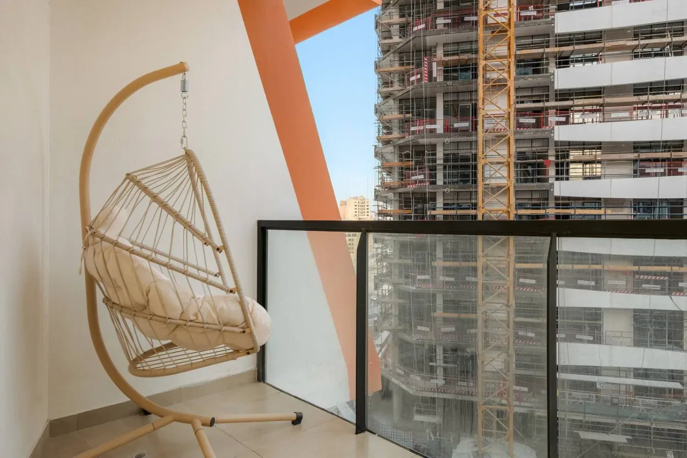 Un balcón con un asiento colgante y una barandilla de vidrio frente a edificios cercanos.