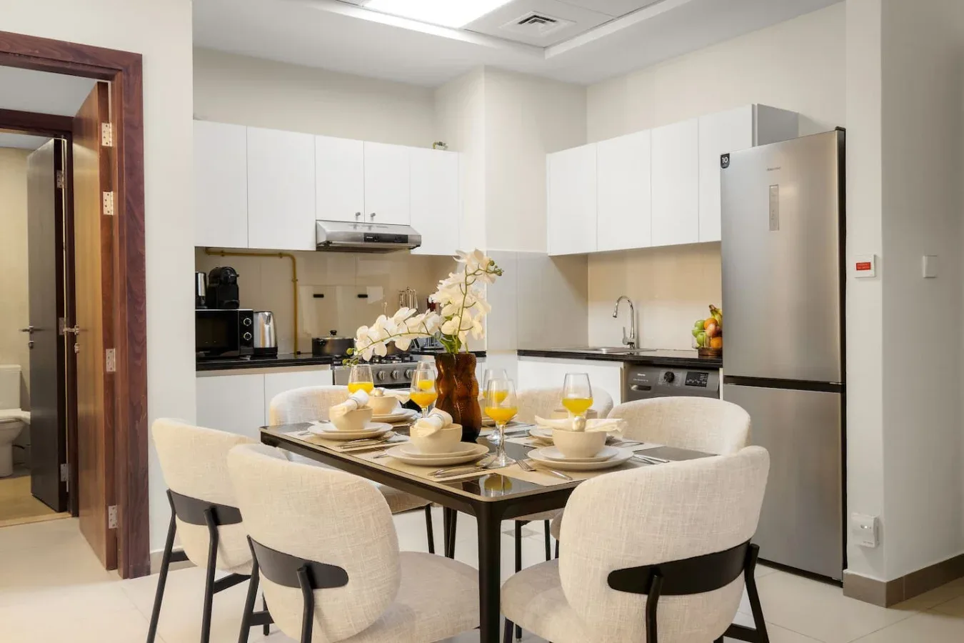 Una mesa de comedor con asientos está frente a una cocina luminosa con gabinetes blancos.