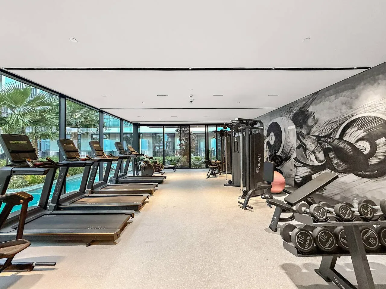 Un gimnasio con cintas de correr junto a las ventanas y pesas junto a la pared.