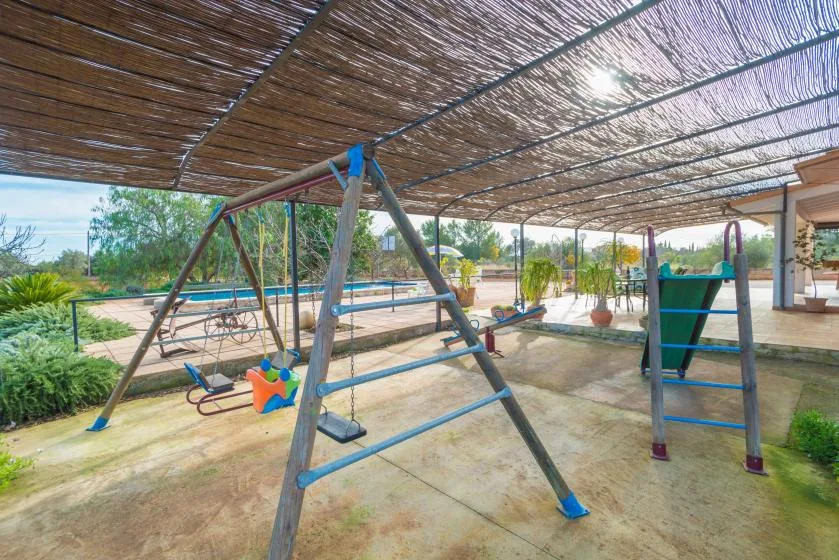 Zona de juegos al aire libre bajo una cubierta de sombra, con columpios y equipo para trepar.