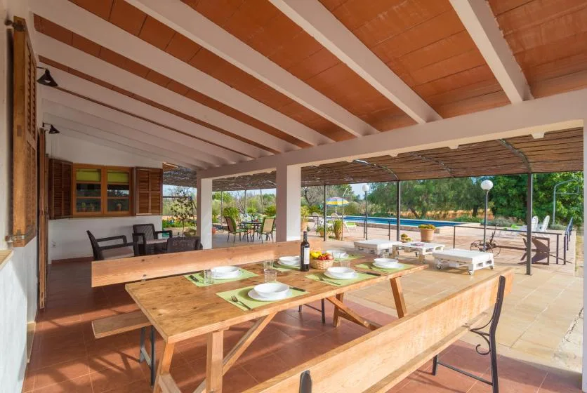 Zona cubierta de comedor al aire libre con mesas de madera junto a una piscina.