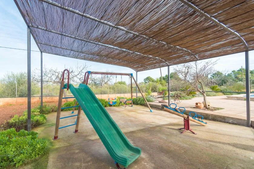 Un parque infantil al aire libre está bajo una cubierta de sombra, con tobogán y columpios.