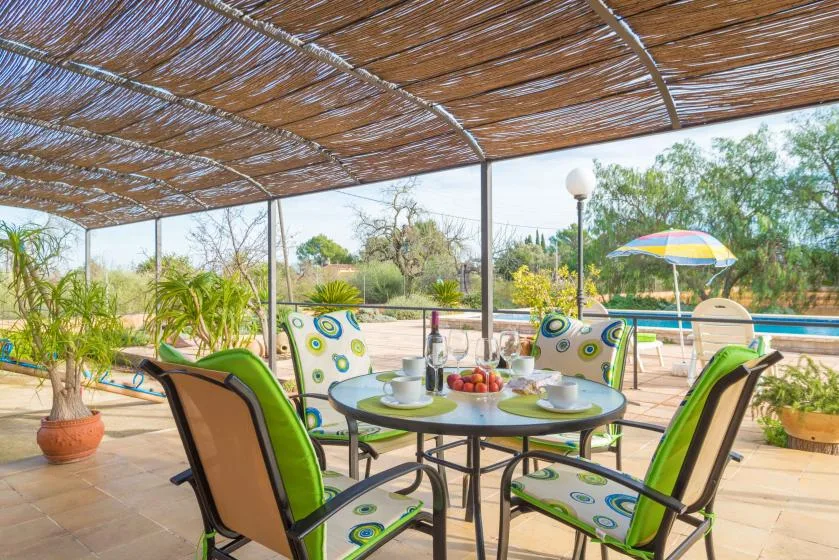 Una zona de comedor al aire libre con sombra da a una piscina y un jardín.