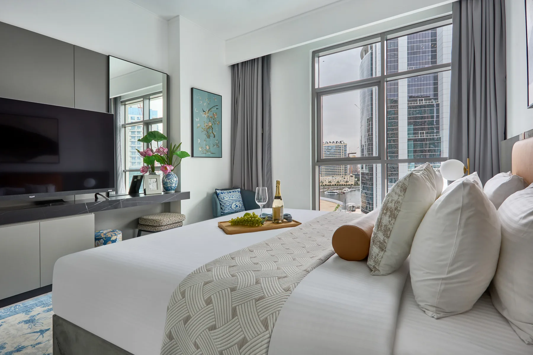 Un dormitorio con una cama bien tendida junto a una gran ventana con vistas a edificios urbanos.
