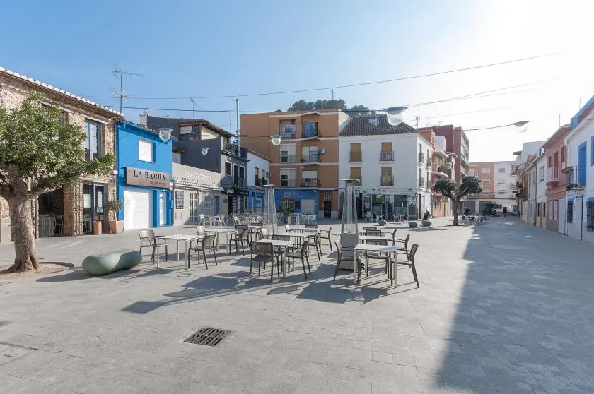 Una plaza pavimentada tiene mesas y asientos al aire libre entre edificios bajos.