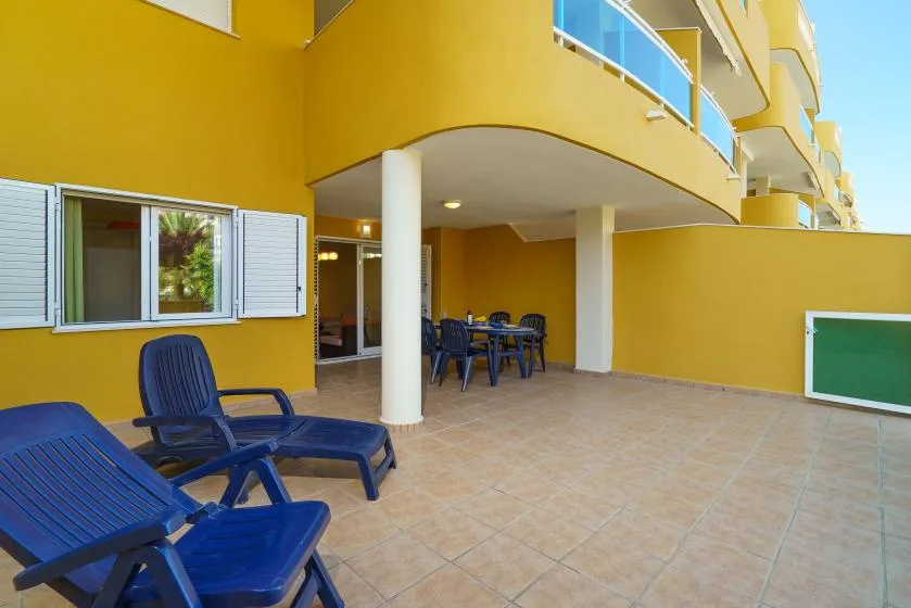 Un patio exterior de baldosas con asientos azules y una mesa de comedor junto a un edificio amarillo.