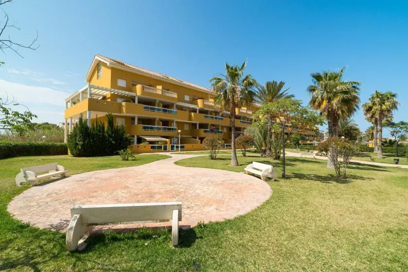 Un edificio amarillo de varias plantas da a un patio ajardinado con palmeras y bancos.
