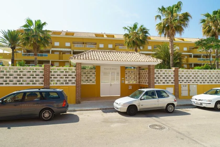 Un edificio de apartamentos amarillo se ve detrás de una entrada cerrada, con coches estacionados en la calle.