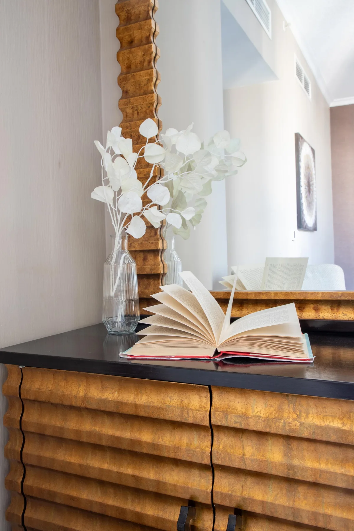 Una sala luminosa con un libro abierto y un jarrón con follaje blanco sobre una consola de madera.