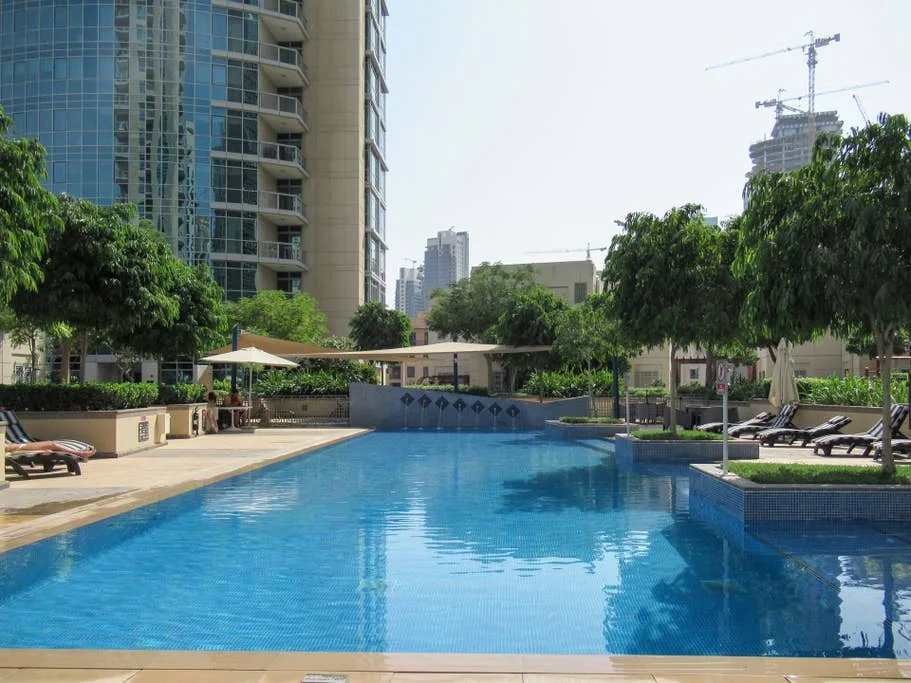 Una piscina junto a un edificio alto, con árboles y asientos alrededor.