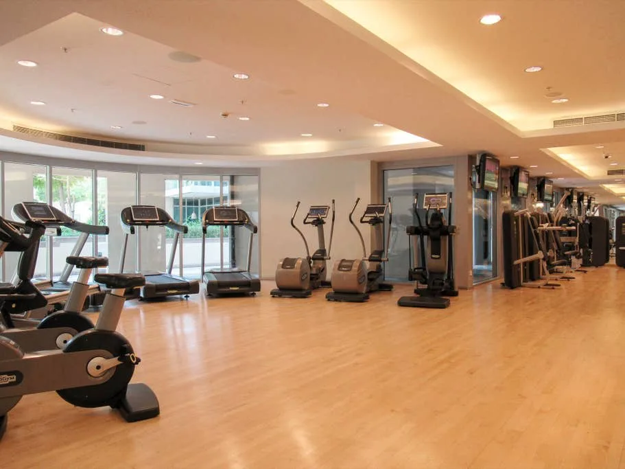 Un gimnasio luminoso con máquinas de cardio junto a las ventanas y equipos de ejercicio por toda la sala.