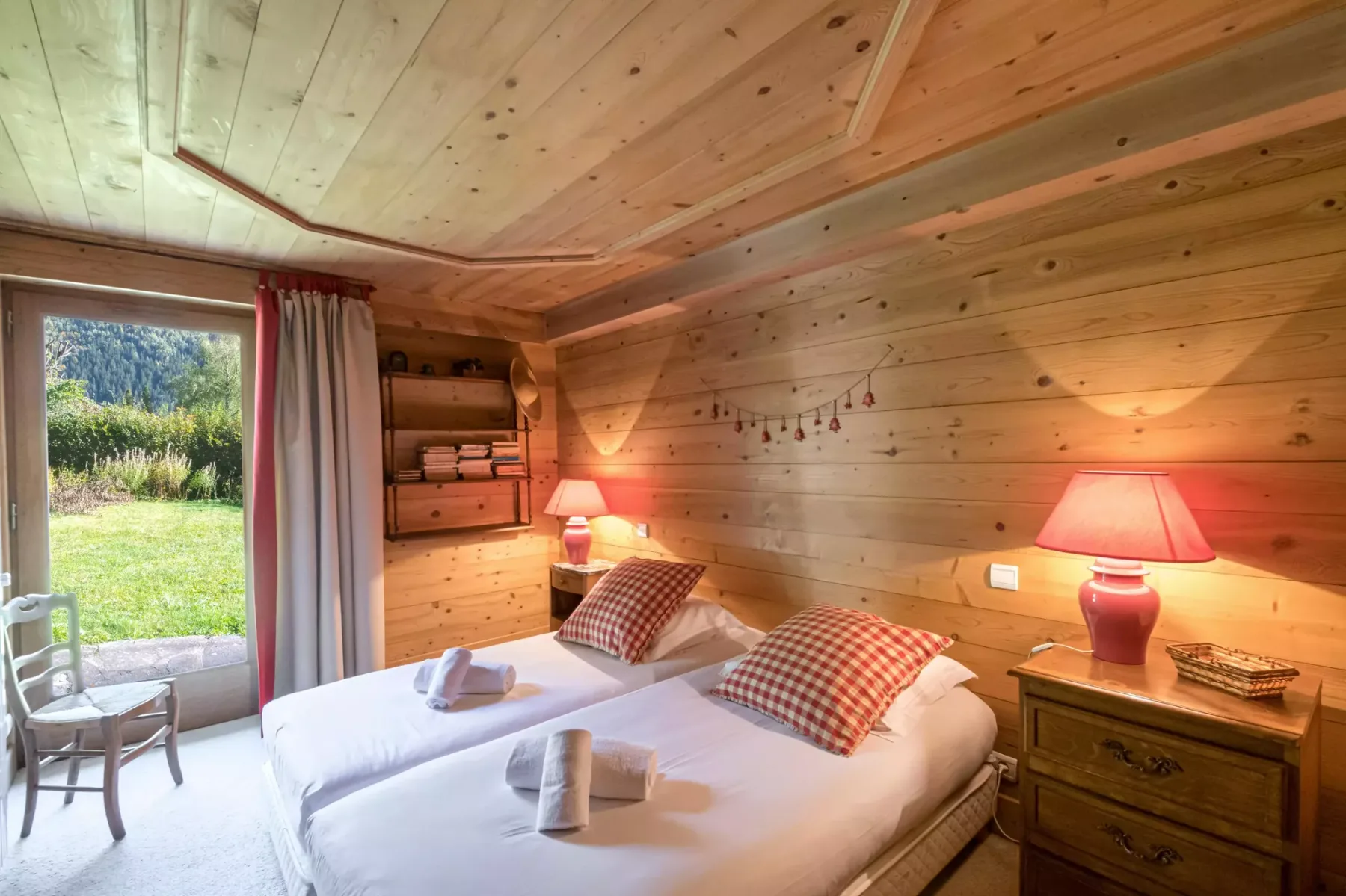 A wood-paneled bedroom with two beds and a window overlooking greenery.