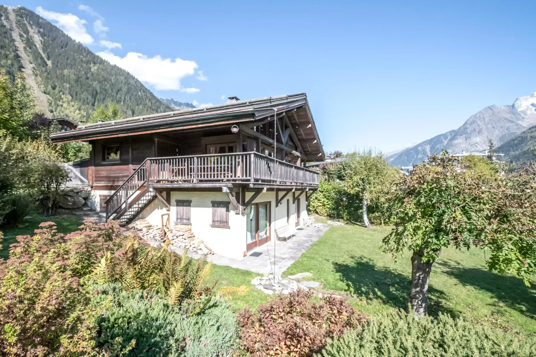 A chalet-style house sits among garden plants with mountain slopes in the background.