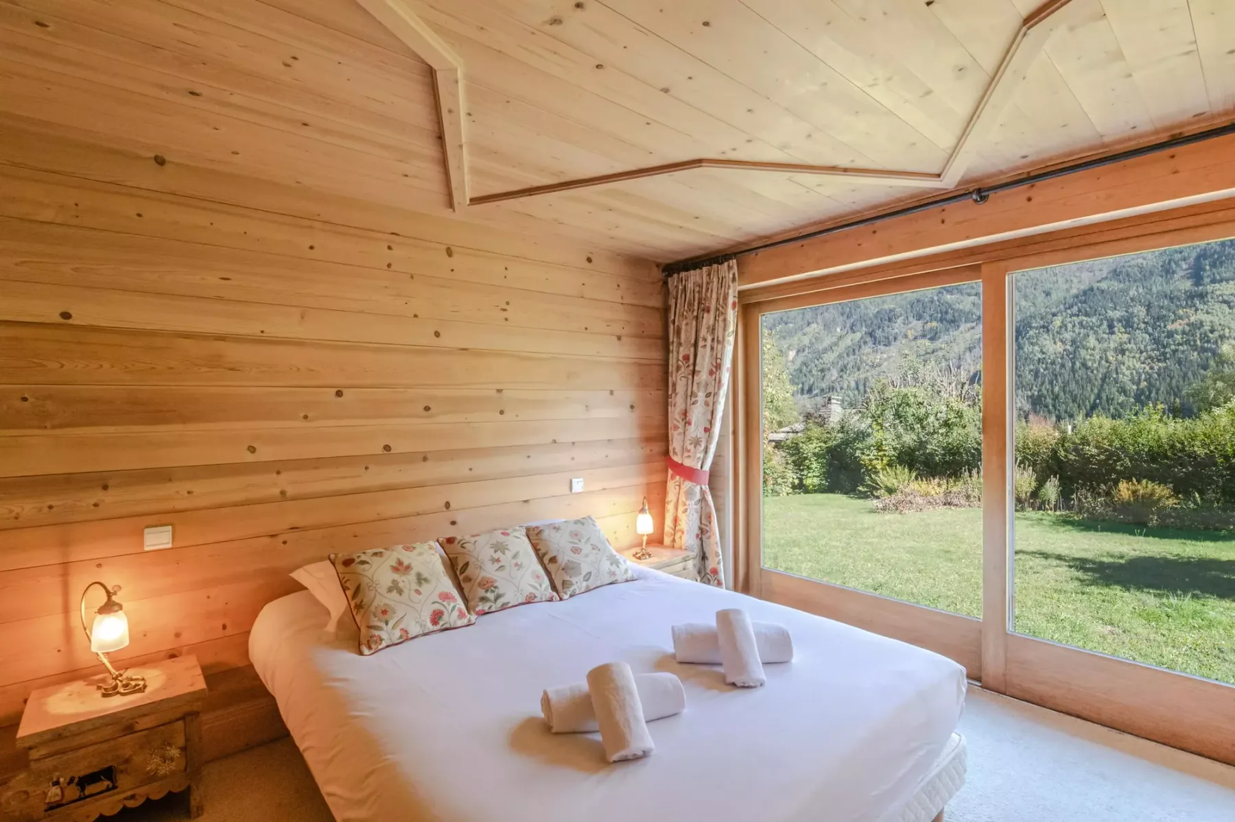 A bedroom with a large bed beside wide windows overlooking a green garden.