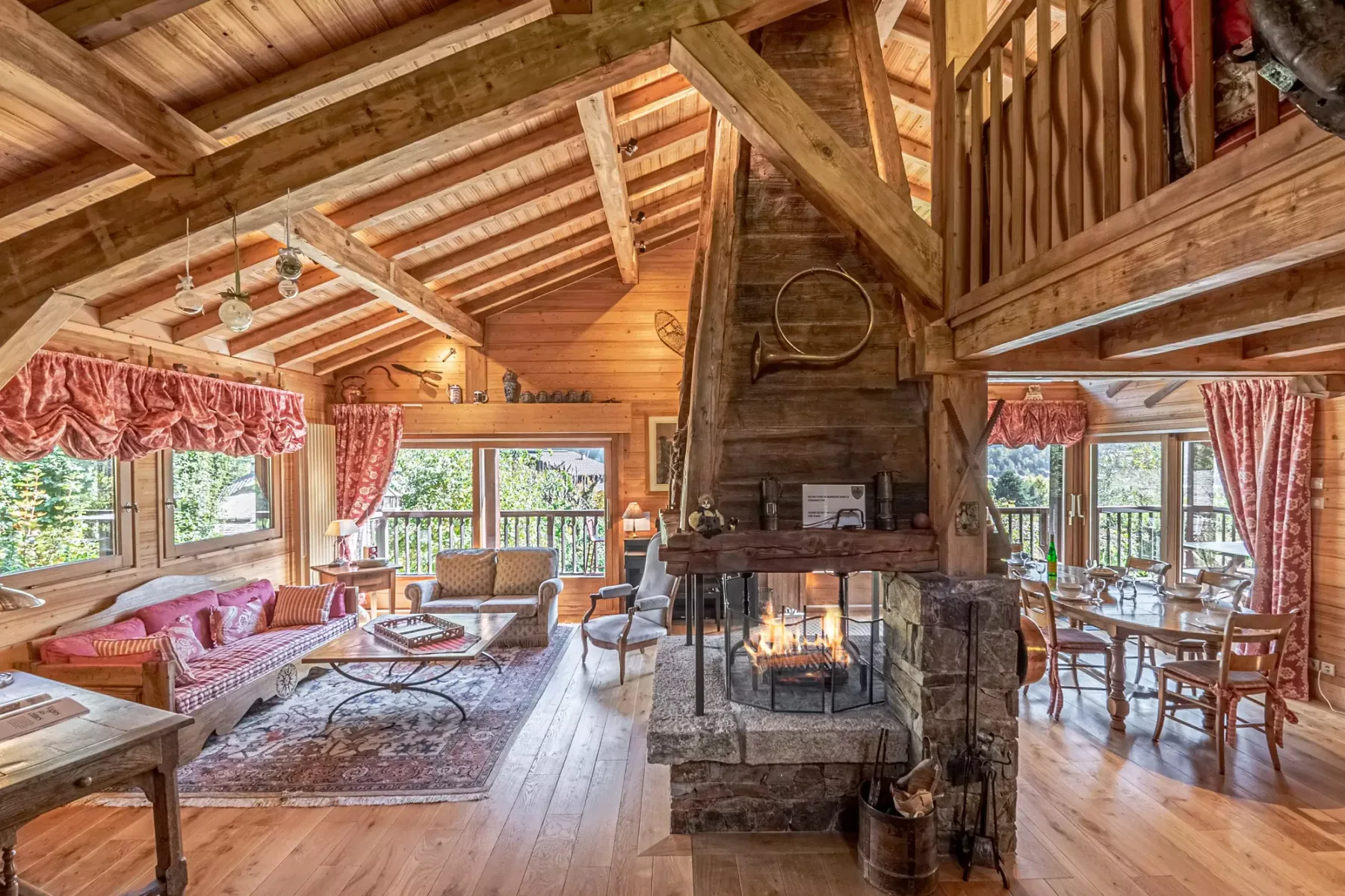 A rustic timber living room with a central stone fireplace and large windows.