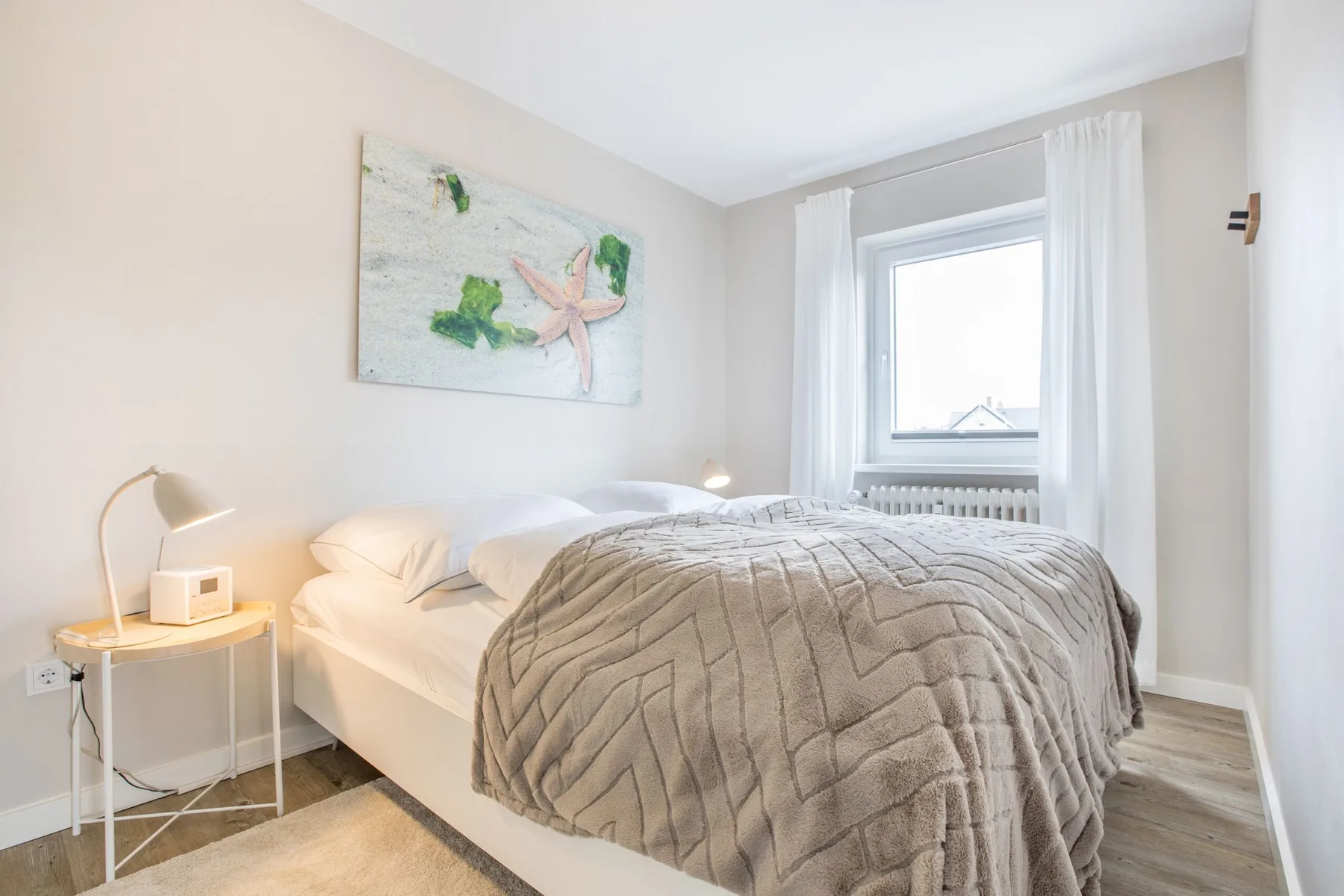 A bright bedroom with a bed beneath a window dressed with white curtains.