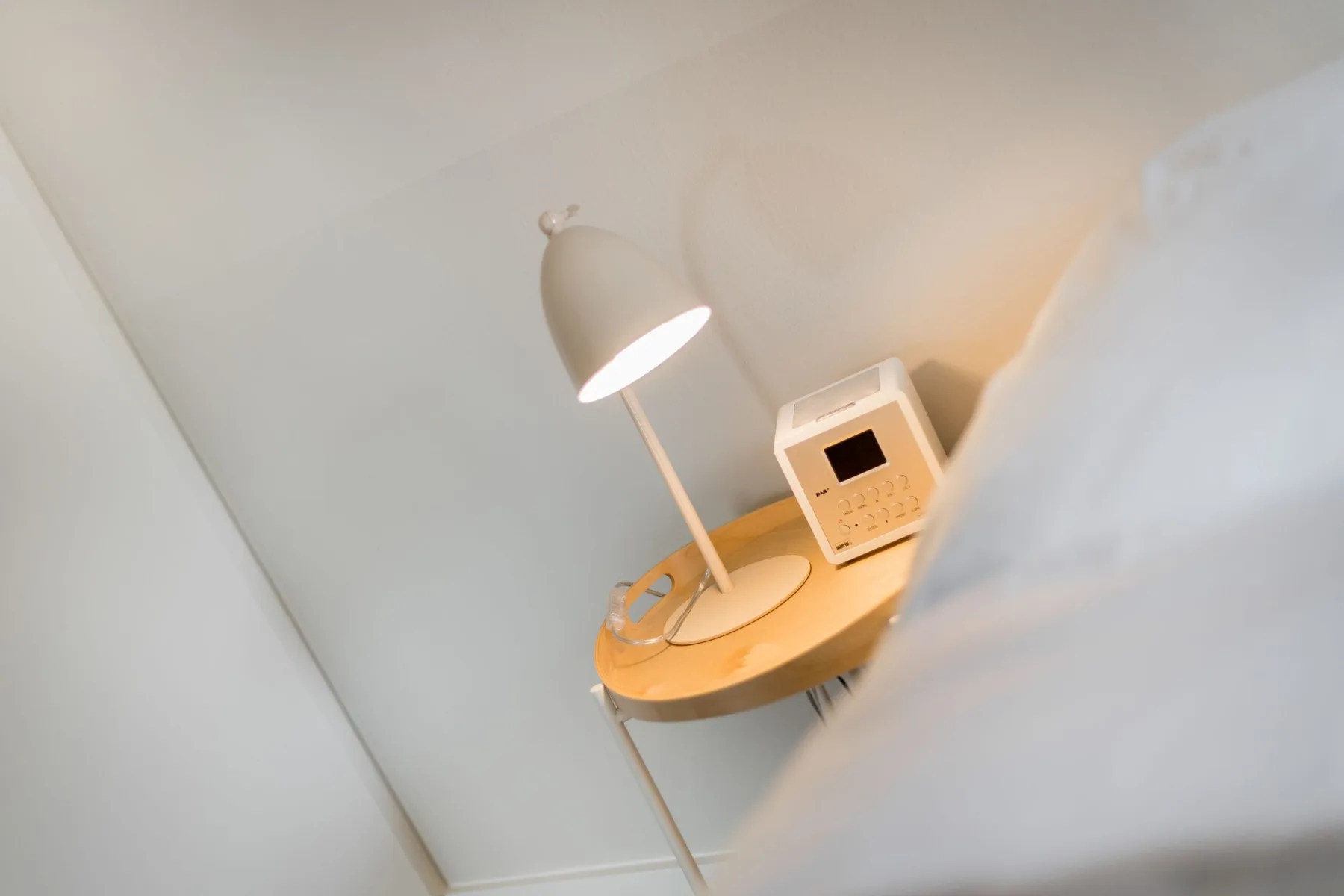 A bedside table holds a lit lamp and a small electronic device.
