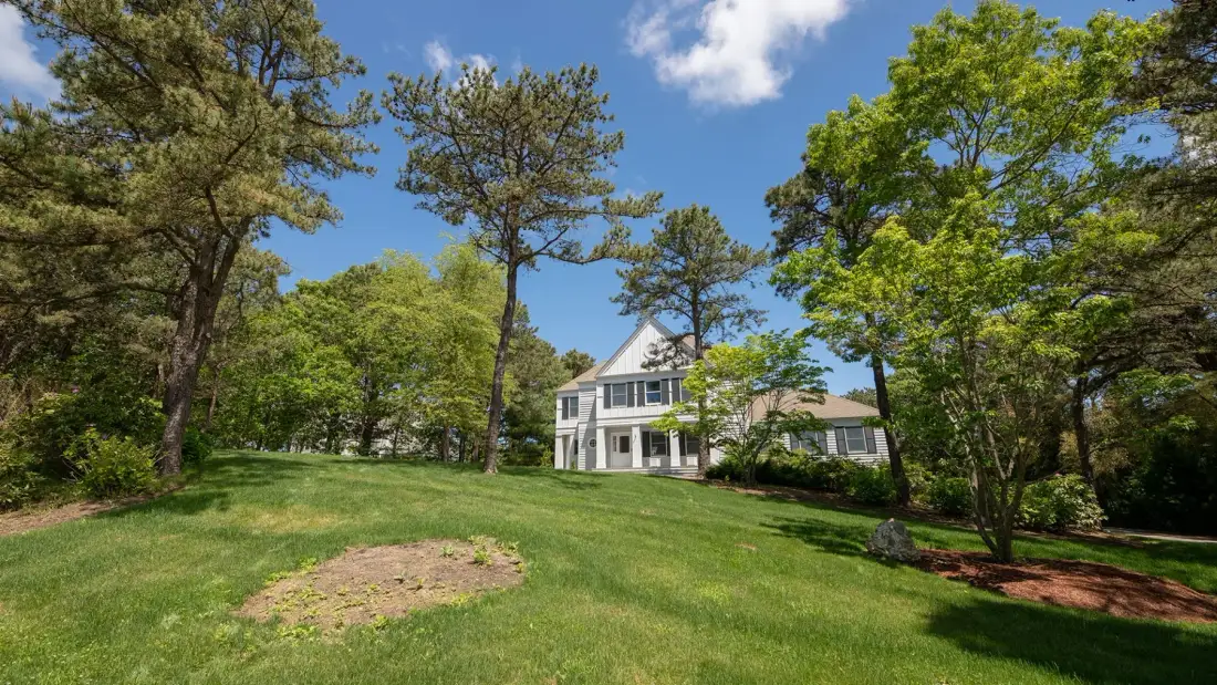 A white house sits beyond a green lawn, surrounded by leafy trees.