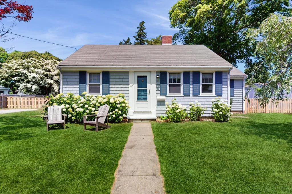 A small house faces a front lawn with a walkway and outdoor seating.